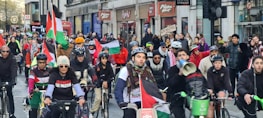 A group of cyclists participating in a protest ride through a city street, carrying Palestinian flags and wearing protest-themed clothing. The crowd is diverse and exhibits solidarity, with some participants holding signs. The street is lined with buildings and storefronts, filled with spectators and more demonstrators.