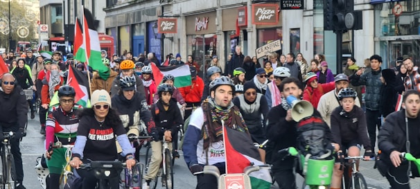 A group of cyclists participating in a protest ride through a city street, carrying Palestinian flags and wearing protest-themed clothing. The crowd is diverse and exhibits solidarity, with some participants holding signs. The street is lined with buildings and storefronts, filled with spectators and more demonstrators.
