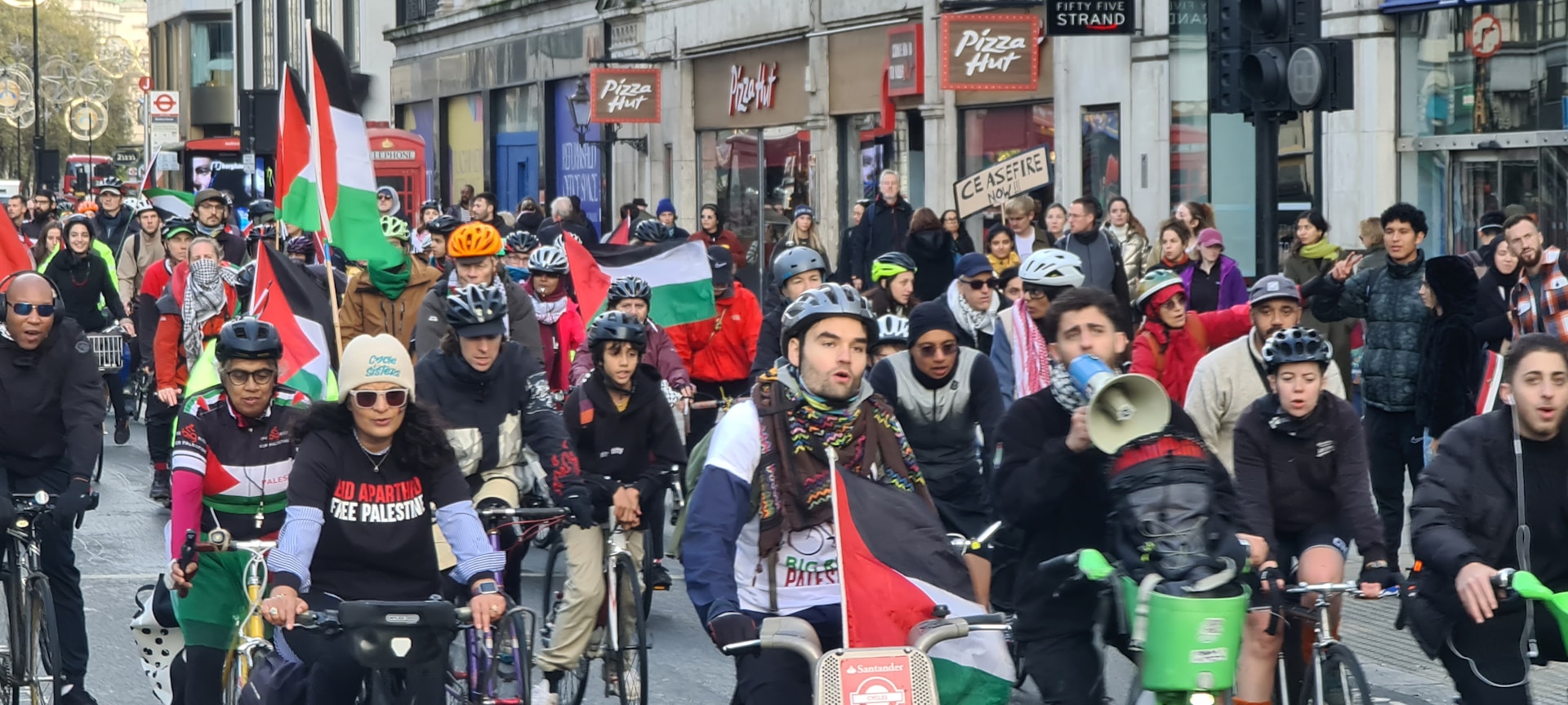 A group of cyclists participating in a protest ride through a city street, carrying Palestinian flags and wearing protest-themed clothing. The crowd is diverse and exhibits solidarity, with some participants holding signs. The street is lined with buildings and storefronts, filled with spectators and more demonstrators.
