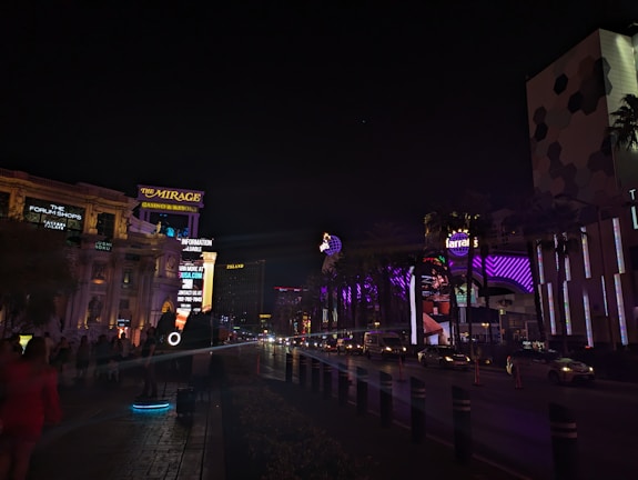 A nighttime urban scene with illuminated signs and buildings. The Mirage Casino & Resort sign is prominently visible, along with other venues and advertisements. There are several cars on the road and people walking along the sidewalk. The area is well-lit with neon and LED lights in various colors.