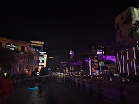 A nighttime urban scene with illuminated signs and buildings. The Mirage Casino & Resort sign is prominently visible, along with other venues and advertisements. There are several cars on the road and people walking along the sidewalk. The area is well-lit with neon and LED lights in various colors.