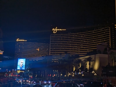 The image features two large hotel buildings with the names 'Encore' and 'Wynn' illuminated in bright lights against a night sky. The buildings display horizontal lines of golden lights, adding to their grandeur. Trees and lampposts are visible near the base of the structures, and a roadway with cars is seen in the foreground. A brightly lit billboard is also visible.
