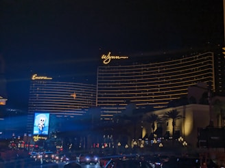 The image features two large hotel buildings with the names 'Encore' and 'Wynn' illuminated in bright lights against a night sky. The buildings display horizontal lines of golden lights, adding to their grandeur. Trees and lampposts are visible near the base of the structures, and a roadway with cars is seen in the foreground. A brightly lit billboard is also visible.