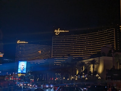 The image features two large hotel buildings with the names 'Encore' and 'Wynn' illuminated in bright lights against a night sky. The buildings display horizontal lines of golden lights, adding to their grandeur. Trees and lampposts are visible near the base of the structures, and a roadway with cars is seen in the foreground. A brightly lit billboard is also visible.