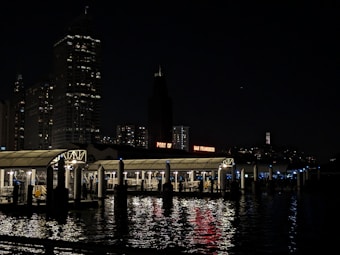 A nighttime scene featuring the illuminated 'Port of San Francisco' sign with buildings towering in the background. Reflections of lights shimmer on the water in the foreground, with a covered pier structure visible.
