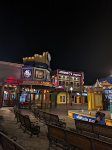 A vibrant night scene showcasing a boardwalk area with various attractions and eateries. Illuminated signs, including advertisements for a pizzeria and a sports grill arcade, are visible. Several benches are positioned along the wooden planks. To the right, a colorful booth and a stage with 'smartwater' signage are featured.