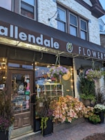 A charming flower shop exterior with a wooden door and a large display window. The storefront is adorned with various potted plants and colorful flowers hanging and arranged at the entrance. The sign above the shop reads 'Allendale Flowers' with decorative elements, and small lights add warmth to the scene.