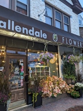A charming flower shop exterior with a wooden door and a large display window. The storefront is adorned with various potted plants and colorful flowers hanging and arranged at the entrance. The sign above the shop reads 'Allendale Flowers' with decorative elements, and small lights add warmth to the scene.