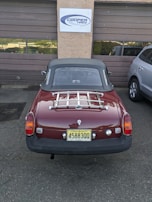 A classic red convertible car with a New Jersey license plate is parked in front of a garage with brown doors. A sign above the garage reads 'Cooper Tires'. The car has a chrome luggage rack on the trunk and is parked next to a modern silver SUV.