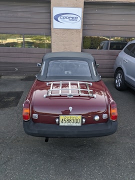 A classic red convertible car with a New Jersey license plate is parked in front of a garage with brown doors. A sign above the garage reads 'Cooper Tires'. The car has a chrome luggage rack on the trunk and is parked next to a modern silver SUV.