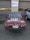 A classic red convertible car with a New Jersey license plate is parked in front of a garage with brown doors. A sign above the garage reads 'Cooper Tires'. The car has a chrome luggage rack on the trunk and is parked next to a modern silver SUV.