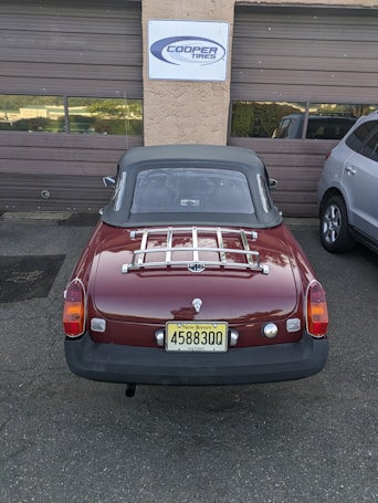 A classic red convertible car with a New Jersey license plate is parked in front of a garage with brown doors. A sign above the garage reads 'Cooper Tires'. The car has a chrome luggage rack on the trunk and is parked next to a modern silver SUV.
