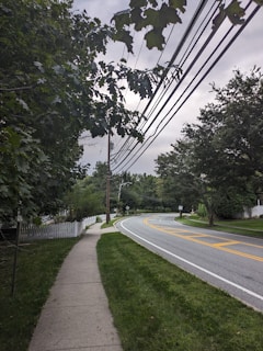 A quiet suburban street with a sidewalk on the left flanked by grass and a white picket fence. Overhead, power lines run parallel to the road, which curves gently to the right. Trees with dense foliage are present on both sides of the street creating a lush, green environment. The sky is overcast, giving the scene a tranquil and calm feel.
