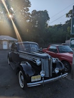 A vintage black car with chrome details is parked outdoors, illuminated by a sunbeam filtering through the trees. The setting includes a small building in the background and another red vehicle partially visible.