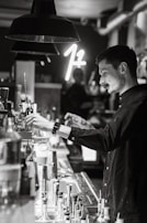 A person is working behind a bar, preparing a drink. The setting is dimly lit, with a modern and stylish atmosphere. Various bar tools and bottles are visible on the counter, and a neon sign glows softly in the background.
