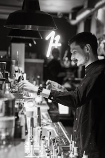 A person is working behind a bar, preparing a drink. The setting is dimly lit, with a modern and stylish atmosphere. Various bar tools and bottles are visible on the counter, and a neon sign glows softly in the background.
