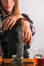 A person is sitting with their hands resting on their knee, wearing a beaded bracelet. The focus on the hands and bracelet creates a serene and relaxed atmosphere, while the background includes out-of-focus objects on a table, possibly cups or small containers.