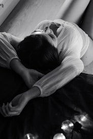 A person is lying down on a surface with their arms stretched back, wearing a light, semi-transparent top. The setting is dimly lit, creating a dramatic play of light and shadow, contributing to a serene and intimate atmosphere.