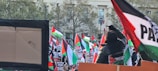 A large group of people holding and waving Palestinian flags, gathered in front of a building with trees in the background. Many signs and banners with various messages are visible, creating a scene of a public demonstration or protest.