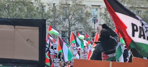 A large group of people holding and waving Palestinian flags, gathered in front of a building with trees in the background. Many signs and banners with various messages are visible, creating a scene of a public demonstration or protest.