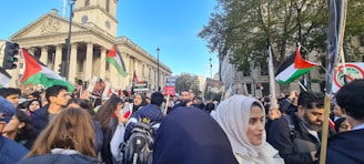 A large group of people are gathered in a city street for a demonstration or protest. Many individuals are holding Palestinian flags and placards with various messages. The scene is set against an urban backdrop with historic architecture and trees.