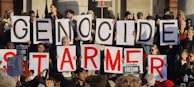 A group of protesters is holding large signs with letters that form a message. The crowd is diverse, with people of various ages and ethnicities. Some individuals are holding smaller signs with messages related to freedom and political themes. The setting appears to be outdoors, possibly near a building with a stone facade.