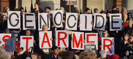 A group of protesters is holding large signs with letters that form a message. The crowd is diverse, with people of various ages and ethnicities. Some individuals are holding smaller signs with messages related to freedom and political themes. The setting appears to be outdoors, possibly near a building with a stone facade.