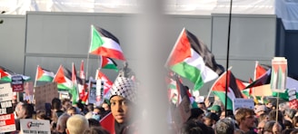 A large crowd of people gathered at a protest, holding Palestinian flags and various signs advocating for Palestinian rights. Many are wearing headscarves and the atmosphere is lively and engaged.