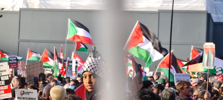 A large crowd of people gathered at a protest, holding Palestinian flags and various signs advocating for Palestinian rights. Many are wearing headscarves and the atmosphere is lively and engaged.