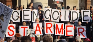 A group of people holding large signs that form the words 'GENOCIDE' and 'STARMER'. Additional signs and placards can be seen, one of them referencing Palestine and another depicting a caricature with the word 'CRIMINAL'. The setting appears to be outdoors with people gathered closely together.