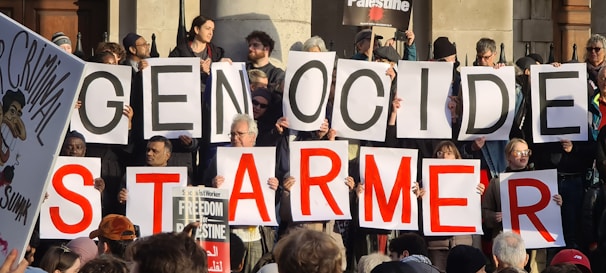 A group of people holding large signs that form the words 'GENOCIDE' and 'STARMER'. Additional signs and placards can be seen, one of them referencing Palestine and another depicting a caricature with the word 'CRIMINAL'. The setting appears to be outdoors with people gathered closely together.