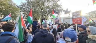 A large crowd gathers in a public square, holding flags and signs in support of Palestine. Many people wear winter clothing, and there is a prominent equestrian statue in the background. The scene is lively, with flags predominantly in red, green, white, and black. Trees with autumn leaves frame the gathering.