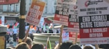 A crowd of people is gathered at a protest, holding up various signs and banners. The signs include statements such as 'END ISRAELI STATE TERROR' and 'STOP THE WAR ON GAZA' and promote a socialist perspective. The photo is taken outdoors, and multiple Palestinian flags are visible. The background includes buildings and a red bus.