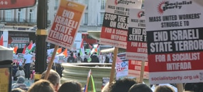 A crowd of people is gathered at a protest, holding up various signs and banners. The signs include statements such as 'END ISRAELI STATE TERROR' and 'STOP THE WAR ON GAZA' and promote a socialist perspective. The photo is taken outdoors, and multiple Palestinian flags are visible. The background includes buildings and a red bus.