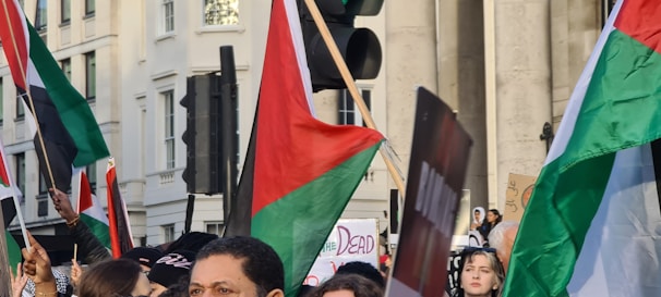 A group of people is gathered, holding flags with red, green, white, and black colors prominently displayed. The setting appears to be an urban environment, possibly during a protest or demonstration. There are signs with text, although specific details are not clear.