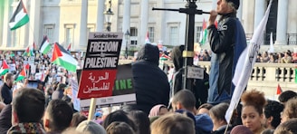 A large crowd of people is gathered in front of a building with columns, holding signs and flags. The predominant flag is the Palestinian flag, and several signs in English and Arabic express support for Palestine. A person stands elevated, speaking to the crowd. The atmosphere is lively and engaged.
