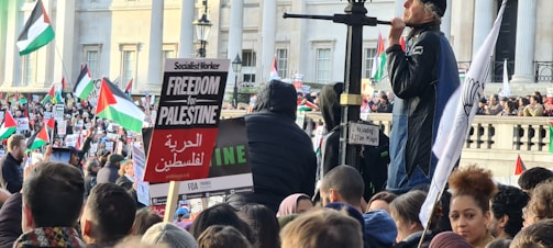 A large crowd of people is gathered in front of a building with columns, holding signs and flags. The predominant flag is the Palestinian flag, and several signs in English and Arabic express support for Palestine. A person stands elevated, speaking to the crowd. The atmosphere is lively and engaged.