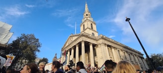 A large crowd gathers in front of a historic church with tall columns and a prominent spire, set against a clear blue sky. Many people are holding signs and flags, indicating the presence of a protest or demonstration. The scene is vibrant with activity and includes a mixture of people from different backgrounds.