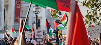 A large gathering of people demonstratively waving flags and holding signs in front of an urban building. The flags feature black, white, green, and red colors, typical of the Palestinian flag. Several people hold signs with political messages, expressing demands for action or change. The scene is lively and appears to be part of a protest or rally. Tall structures and a streetlamp are visible in the background.