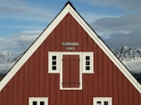 A red wooden building with white trim and a sign reading 'Langabúð 1790' on the facade. It sits against a backdrop of snow-capped mountains under a partly cloudy sky.