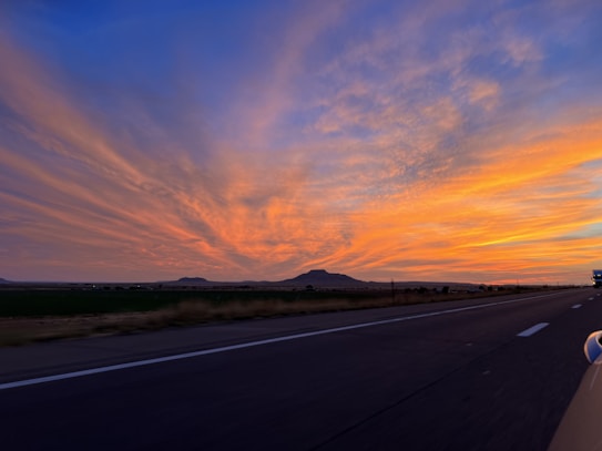 A wide-open road stretches toward the horizon, flanked by open fields and distant hills. The sky is filled with vibrant colors of the setting sun, predominantly shades of orange, pink, and blue. Wispy clouds spread out, creating a dramatic and picturesque sunset scene.