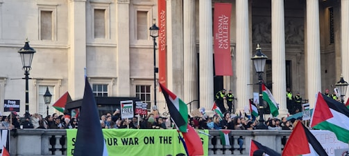 A large crowd of protesters is gathered outside a historic building with columns, holding Palestinian flags and banners. Signs reading 'Free Palestine' and other messages are visible. The building has a sign indicating it is the National Gallery. There are police officers present, and several large street lamps line the area.