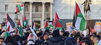A large crowd of people is gathered, holding flags and protest signs. The flags have a red, white, black, and green color scheme. The setting seems to be an urban area with historical architecture in the background, and a statue of a horse and rider is visible. A cluster of balloons in red, green, white, and black are floating above the crowd.