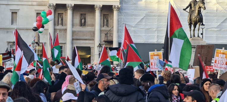 A large crowd of people is gathered, holding flags and protest signs. The flags have a red, white, black, and green color scheme. The setting seems to be an urban area with historical architecture in the background, and a statue of a horse and rider is visible. A cluster of balloons in red, green, white, and black are floating above the crowd.