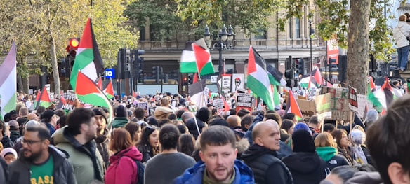 A large crowd gathers in a city setting under trees, with many people holding Palestinian flags and signs advocating for Palestine. The scene appears lively and densely packed, with a mix of different people in various clothing and jackets.