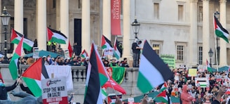 A crowded scene with people holding Palestinian flags and protest signs in front of a large building with columns. The signs have messages related to stopping the war on Gaza and other related statements. The crowd is densely packed, showing an organized protest or demonstration.