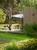 A serene wooden deck surrounded by lush greenery, featuring a small outdoor seating area with a folding table and chairs. A large white umbrella provides shade, and a nearby tree adds a natural touch. A portable grill is present on the deck, and a wooden structure, possibly a house or cabin, is partially visible. The backdrop includes a forest with tall, dense trees.