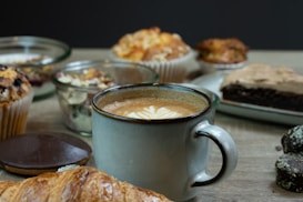 A mug of cappuccino with latte art is surrounded by an assortment of pastries including a croissant, muffins, and brownies. The setting appears cozy, with the pastries and coffee arranged on a wooden surface.