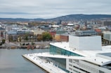 A modern waterfront building with a geometric design stands prominently in the lower right, featuring a sloped roof with people walking on it. The structure is positioned near a body of water, and in the background, a cityscape with various buildings, trees, and hills is visible under a cloudy sky.