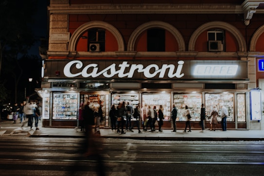 A group of people stand outside a well-lit shop named Castroni Caff&egrave;. The storefront features large windows displaying various products and is flanked by old architectural elements. The scene occurs on a dimly lit street, with blurred pedestrians indicating movement.